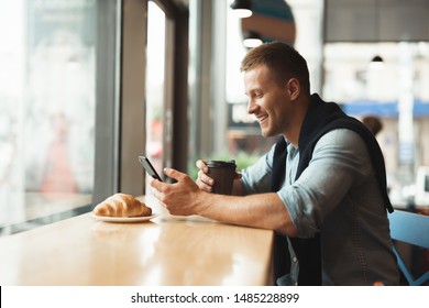 young handsome man smiling having his croissant and drinking hot coffee for lunch in the cafe while watching video in smartphone - Powered by Shutterstock