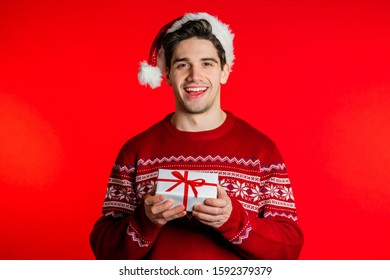 Young Handsome Man Smiling And Holding Gift Box On Red Studio Background. Guy In Santa Hat And Ugly Christmas Sweater. New Year Mood.