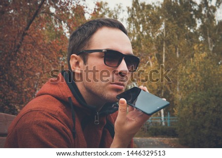 Young handsome man standing in park in summer, using a cell phone to listen to recorded voice message or audio. guy speaks into the microphone to record a voice message. Recording on dictaphone.