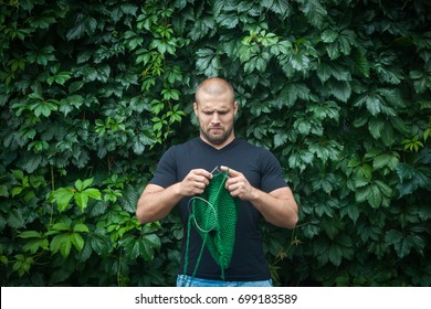 A Young Handsome Man Stands On A Background Of A Green Wall Of Real And Grapes And Knits A Green Sweater With The Knitting Needles Of Their Natural Woolen Threads. A Man Learn To Knit  Needles Sweater