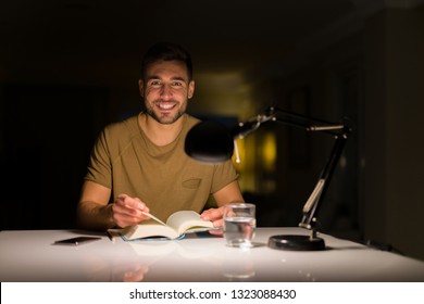 Young Handsome Man Studying At Home, Reading A Book At Night. Smiling Confident At The Camera