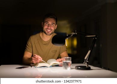 Young Handsome Man Studying At Home, Reading A Book At Night. Smiling Confident At The Camera