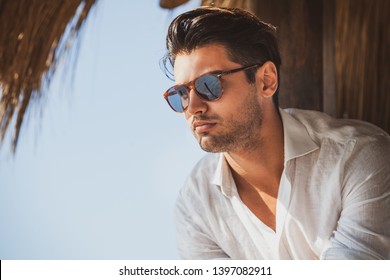 Young and handsome man with sunglasses looking. Warm summer lights, he wears a white shirt. Light short beard. Fashionable hair.