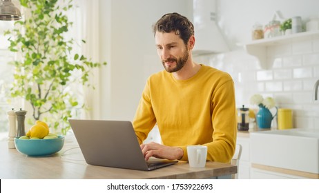 Young Handsome Man Using Laptop Computer And Drinking Morning Cup Of Coffee Or Tea While Wearing A Yellow Pullover. Curly-hared Male Sitting In A Modern Sunny Kitchen. Freelancer Working From Home.