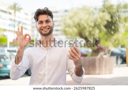 Young handsome man using mobile phone at outdoors showing ok sign with fingers