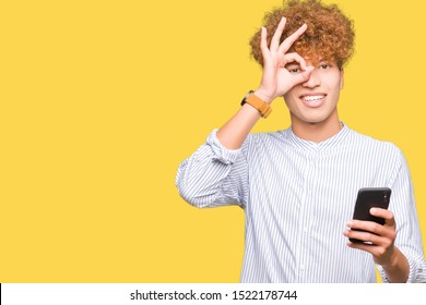Young handsome man using smartphone with happy face smiling doing ok sign with hand on eye looking through fingers