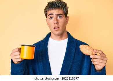 Young Handsome Man Wearing Bathrobe Eating Breakfast Holding Coffee And Croissant In Shock Face, Looking Skeptical And Sarcastic, Surprised With Open Mouth 