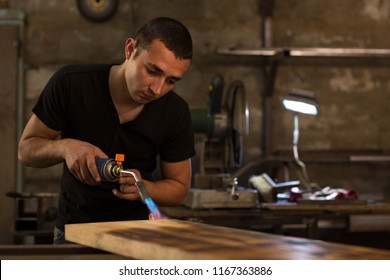 Young handsome man, wearing in black shirt, burning wooden board for displaying textures with petrol blowtorch ignition at factory. Male employee looking at board carefully for producing best pattern. - Powered by Shutterstock