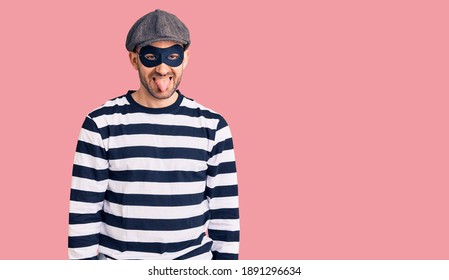 Young Handsome Man Wearing Burglar Mask Sticking Tongue Out Happy With Funny Expression. Emotion Concept. 