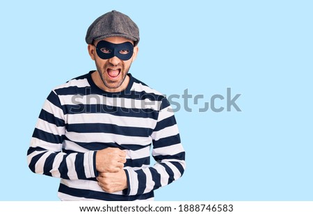 Young handsome man wearing burglar mask smiling and laughing hard out loud because funny crazy joke with hands on body. 
