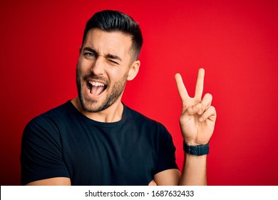 Young Handsome Man Wearing Casual Black T-shirt Standing Over Isolated Red Background Smiling With Happy Face Winking At The Camera Doing Victory Sign. Number Two.