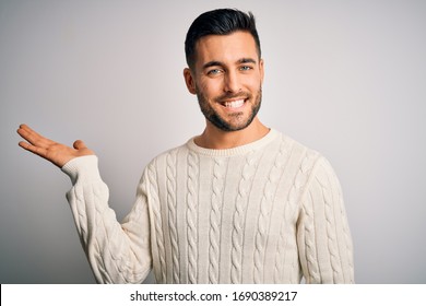 Young handsome man wearing casual sweater standing over isolated white background smiling cheerful presenting and pointing with palm of hand looking at the camera.