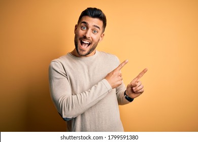 Young handsome man wearing casual sweater standing over isolated yellow background smiling and looking at the camera pointing with two hands and fingers to the side.