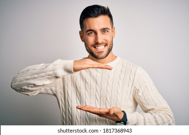 Young handsome man wearing casual sweater standing over isolated white background gesturing with hands showing big and large size sign, measure symbol. Smiling looking at the camera. Measuring.