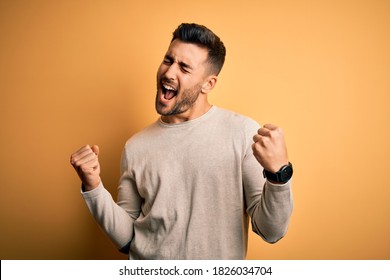 Young handsome man wearing casual sweater standing over isolated yellow background very happy and excited doing winner gesture with arms raised, smiling and screaming for success. Celebration concept. - Powered by Shutterstock