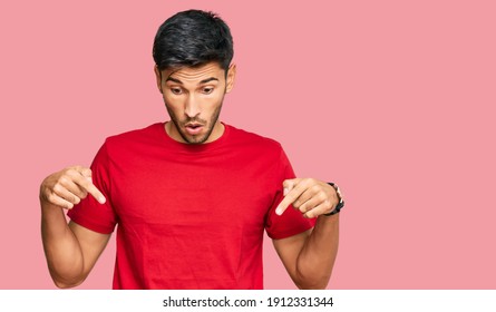 Young handsome man wearing casual red tshirt pointing down with fingers showing advertisement, surprised face and open mouth 
