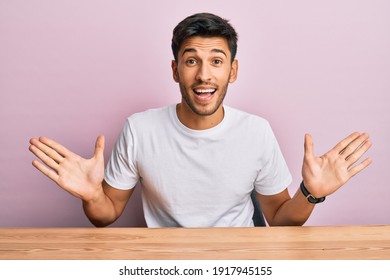 Young Handsome Man Wearing Casual White T-shirt Sitting On The Table Celebrating Victory With Happy Smile And Winner Expression With Raised Hands 