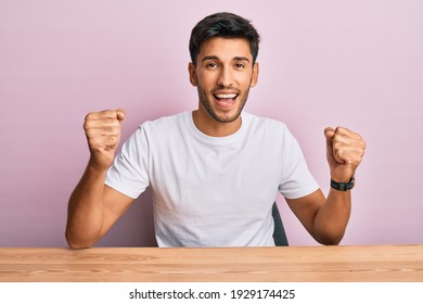 Young Handsome Man Wearing Casual White T-shirt Sitting On The Table Screaming Proud, Celebrating Victory And Success Very Excited With Raised Arms 