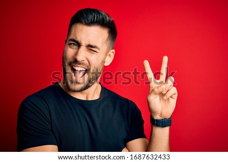 Young handsome man wearing casual black t-shirt standing over isolated red background smiling with happy face winking at the camera doing victory sign. Number two.