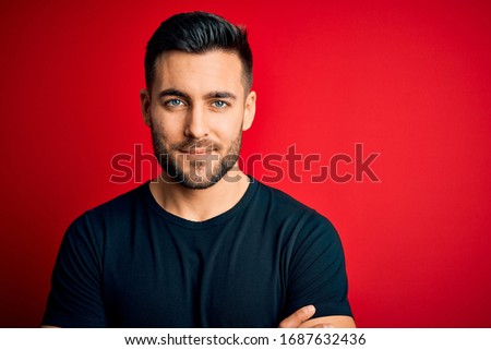Young handsome man wearing casual black t-shirt standing over isolated red background happy face smiling with crossed arms looking at the camera. Positive person.