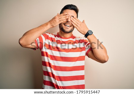 Young handsome man wearing casual striped t-shirt standing over isolated white background covering eyes with hands smiling cheerful and funny. Blind concept.