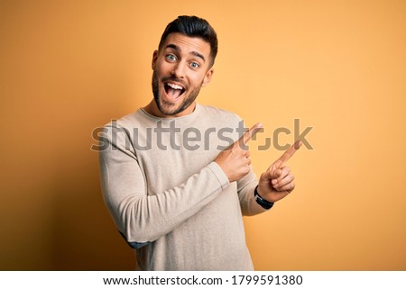 Young handsome man wearing casual sweater standing over isolated yellow background smiling and looking at the camera pointing with two hands and fingers to the side.