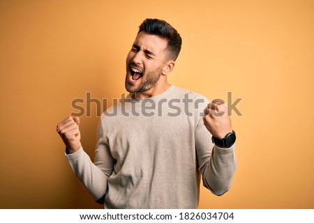 Young handsome man wearing casual sweater standing over isolated yellow background very happy and excited doing winner gesture with arms raised, smiling and screaming for success. Celebration concept. Foto d'archivio © 