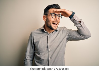 Young handsome man wearing elegant shirt and glasses over isolated white background very happy and smiling looking far away with hand over head. Searching concept.