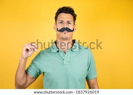 Young handsome man wearing green casual t-shirt over isolated yellow background happy with mustache mask