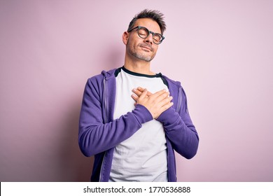 Junge gut aussehende Mann mit violettem Sweatshirt und Brille auf rosafarbenem Hintergrund, lächelnd mit Händen auf der Brust mit geschlossenen Augen und dankbare Geste auf dem Gesicht. Gesundheitskonzept.