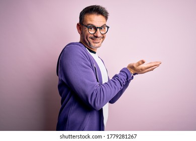 Junge gut aussehende Mann mit violettem Sweatshirt und Brillen auf rosafarbenem Hintergrund, die neben den Handflächen offene Palmen zeigen, die Kopienraum zeigen, und die Werbung, die fröhlich lächelt