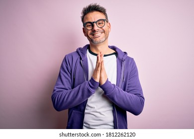 Der junge gut aussehende Mann mit violettem Sweatshirt und Brille auf rosafarbenem Hintergrund betet mit Händen zusammen und bittet um Verzeihung. Er lächelt zuversichtlich.