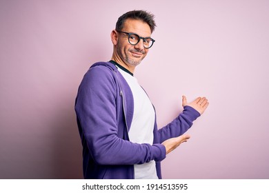 Junge gut aussehende Mann mit violettem Sweatshirt und Brille auf rosafarbenem Hintergrund Einladung, mit offener Hand ins Lächeln der Natur einzutreten