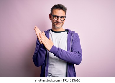 Junge gut aussehende Mann mit violettem Sweatshirt und Brillen auf rosafarbenem Hintergrund, die klatschen und glücklich und fröhlich applaudieren und stolze Hände zusammenlächeln
