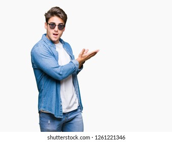 Junge, gut aussehende Mann mit Sonnenbrille auf isoliertem Hintergrund Mit der Hand und der offenen Handfläche zur Seite springen, glücklich und zuversichtlich präsentieren und lächeln