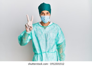 Young handsome man wearing surgeon uniform and medical mask showing and pointing up with fingers number two while smiling confident and happy. 