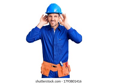 Young Handsome Man Wearing Worker Uniform And Hardhat Trying To Hear Both Hands On Ear Gesture, Curious For Gossip. Hearing Problem, Deaf 