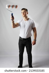 Young handsome man in a white T-shirt and black jeans painting a surface in front of himself with paint roller against a grey background