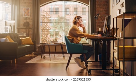 Young Handsome Man Working from Home on Desktop Computer in Sunny Stylish Loft Apartment. Creative Designer Wearing Cozy Yellow Sweater and Headphones. Urban City View from Big Window.