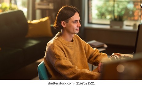 Young Handsome Man Working from Home on Desktop Computer in Sunny Stylish Loft Apartment. Creative Designer Wearing Cozy Yellow Sweater and Headphones. Urban City View from Big Window. Static Shot.