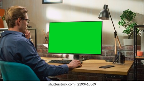 Young Handsome Man Working from Home on Desktop Computer with Green Screen Mock Up Display. Creative Male Checking Social Media, Browsing Internet. Living Room in Bright Loft Apartment. - Powered by Shutterstock