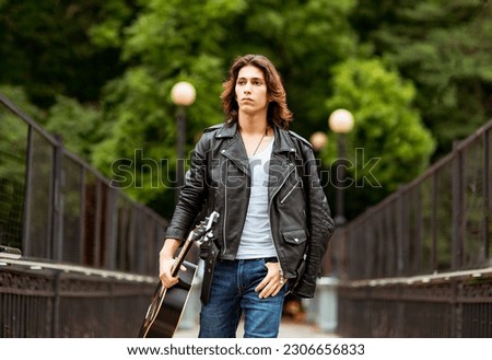 A young, handsome man,brunette with long hair,musician,walks around the city with a guitar, in jeans and a leather jacket