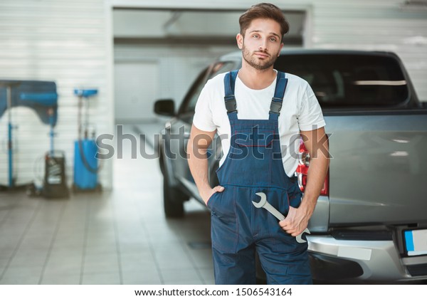 young handsome mechanic wearing uniform with one\
hand in pocket and with spanner in another hand stands in car\
service center