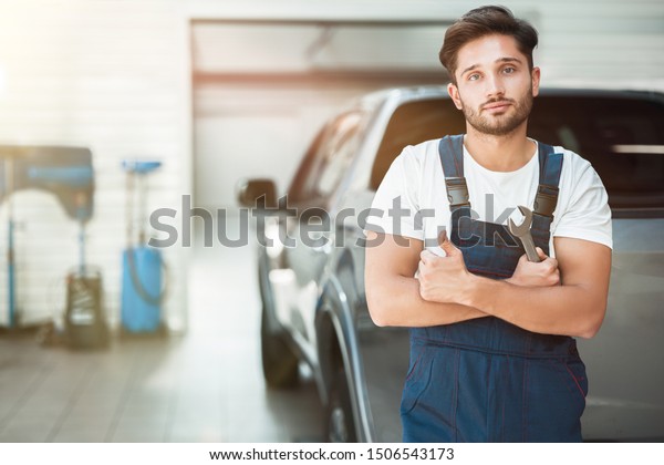 young handsome mechanic\
wearing uniform holding spanner stands in car service center\
showing like sign