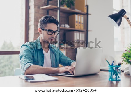 Young handsome minded man in jeans shirt and spectacles sitting at the table and typink a letter to his  acquaintance