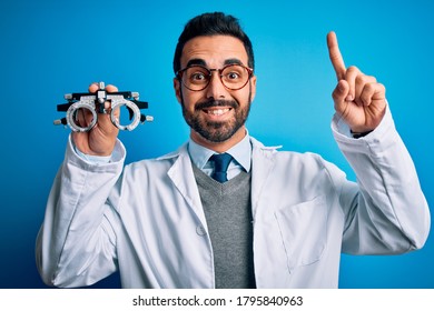 Young Handsome Optical Man With Beard Holding Optometry Glasses Over Blue Background Smiling Amazed And Surprised And Pointing Up With Fingers And Raised Arms.