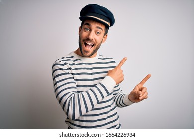 Young handsome sailor man with beard wearing navy striped uniform and captain hat smiling and looking at the camera pointing with two hands and fingers to the side.
