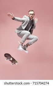 Young handsome serious man with glasses, brown hair and beard, wearing light grey suit and sneakers, jumping with the skateboard and flying in the air on light pink background 