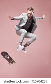 Young handsome serious man with glasses, brown hair and beard, wearing light grey suit and sneakers, jumping with the skateboard and flying in the air on light pink background  - Shutterstock ID 715704283