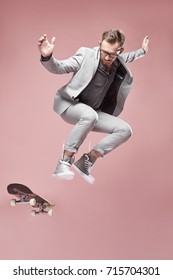 Young handsome serious man with glasses, brown hair and beard, wearing light grey suit and sneakers, jumping with the skateboard and flying on light pink background 